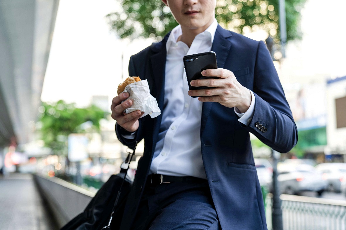 Man standing on the street eating burger while sending email on his phone