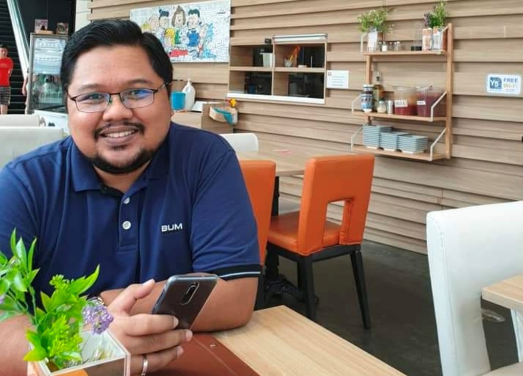 Mr Mohamad Syahid Bin Arif, man holding a phone and seated at a table with a potted plant